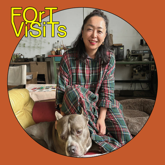 A smiling woman sits indoors with a gray pitbull-type dog on a couch, wearing a green, red, and blue plaid shirt and matching pants. The photo is framed in a circular border with an orange background and the text 'FOR VISITS' in yellow at the top left.