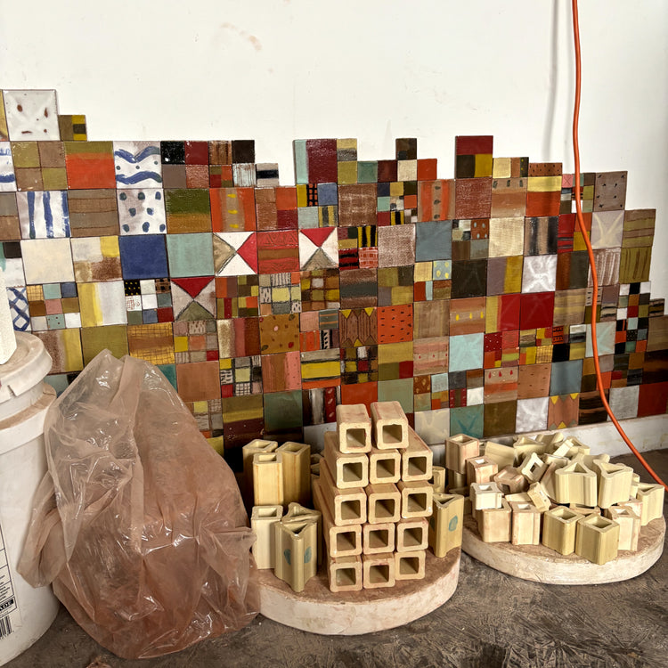 A handmade mosaic tile wall featuring a patchwork of colorful square tiles, with stacks of unfinished light wood blocks and square tiles displayed on round pedestals in front, set against a plain white wall.