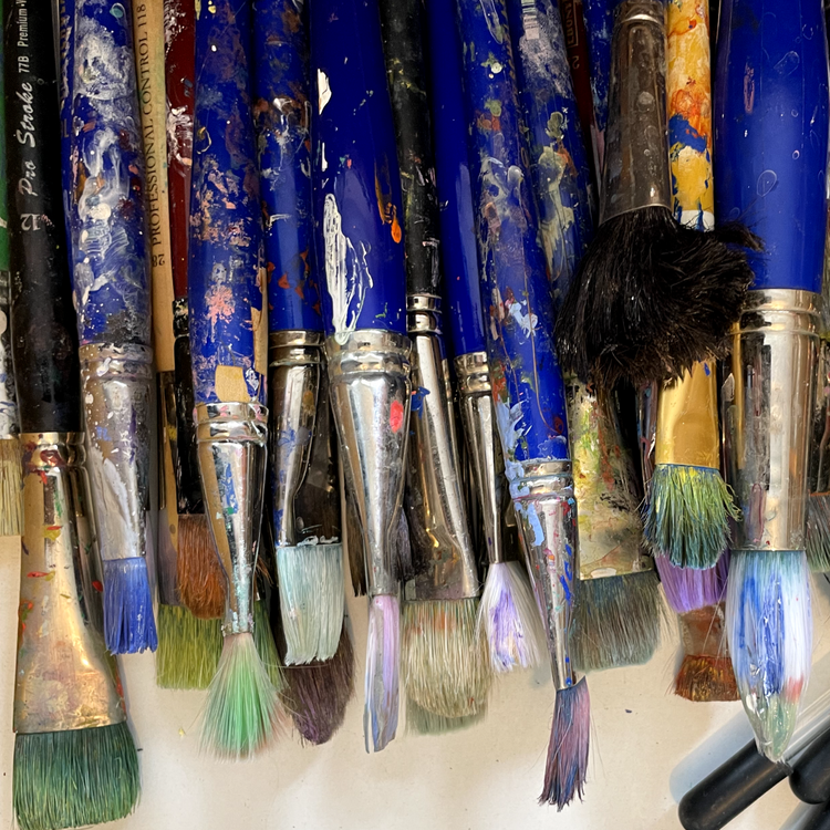 A close-up arrangement of paint brushes with blue handles and silver metal ferrules, many visibly stained with multicolored paint, hanging against a light background.