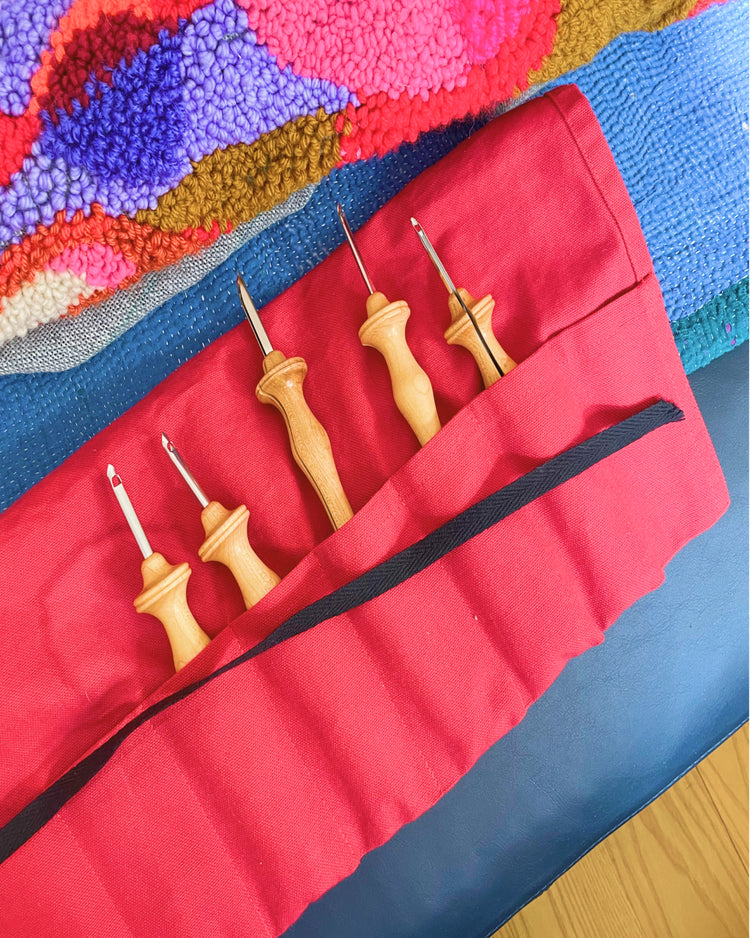 A red fabric tool roll holding five hand tools with light wood handles and metal tips, photographed on a blue surface with a colorful textured rug in the background.