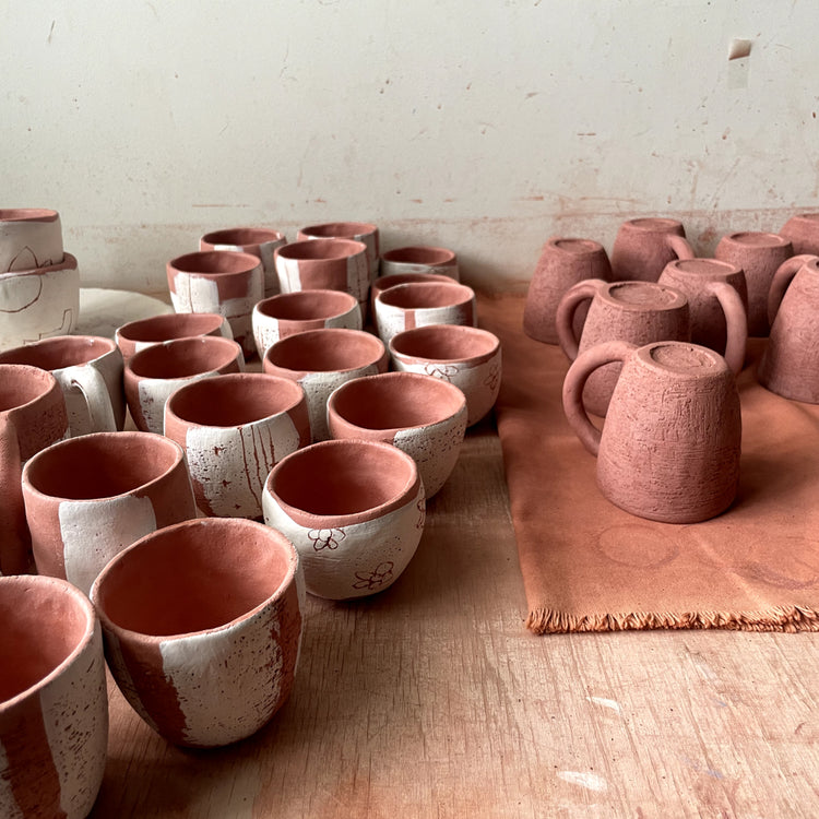 A group of handmade terracotta ceramic mugs and cups arranged on a wooden table in a pottery studio setting, with some pieces resting on a brown paper sheet against a light-colored wall.