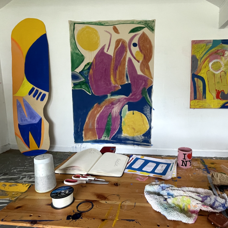 An artist's studio workspace with three colorful abstract paintings hanging on a white wall above a wooden worktable covered with art supplies, including a notebook, paintbrushes, a spray can, and a paint-splattered towel.