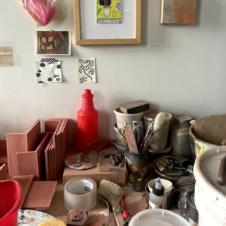 A cluttered artist studio or craft workspace with terracotta clay blocks, paint cans, brushes, tools, tape, and framed artwork on a wall.