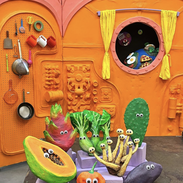 A colorful children's play kitchen scene with a bright orange textured kitchen wall, yellow curtains, a round window, and various sculpted vegetables and fruits with cartoon eyes, including a papaya, broccoli, cactus, eggplant, and pumpkin.