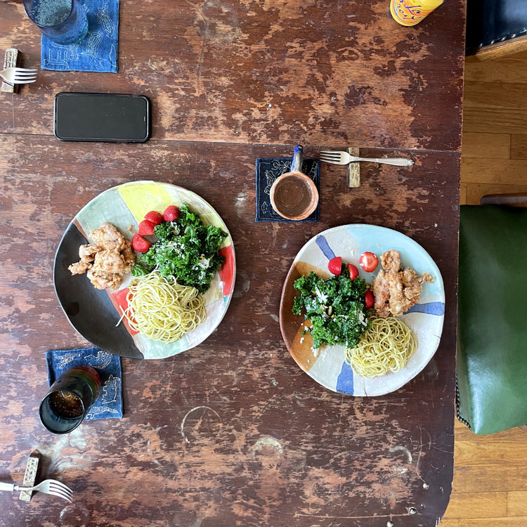 Two colorful ceramic plates of pasta salad on a rustic wooden table, surrounded by cutlery, a smartphone, and a small cup.