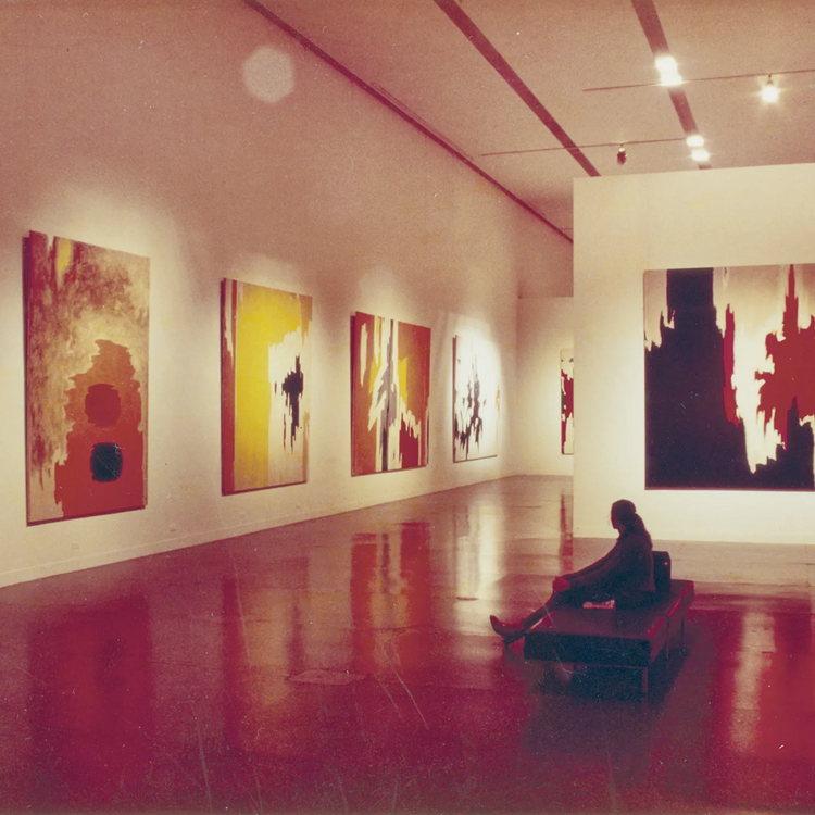 An abstract art gallery scene with several large paintings in red, yellow, black, and white tones hanging on white walls. A person sits on a bench in the foreground on a glossy red floor.