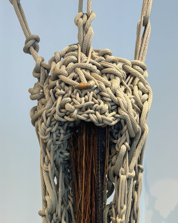 A close-up of a large woven rope basket or net in natural off-white and gray tones, suspended against a pale blue sky. A dark wooden pole runs through the center, and the top of the basket is attached to hanging ropes.