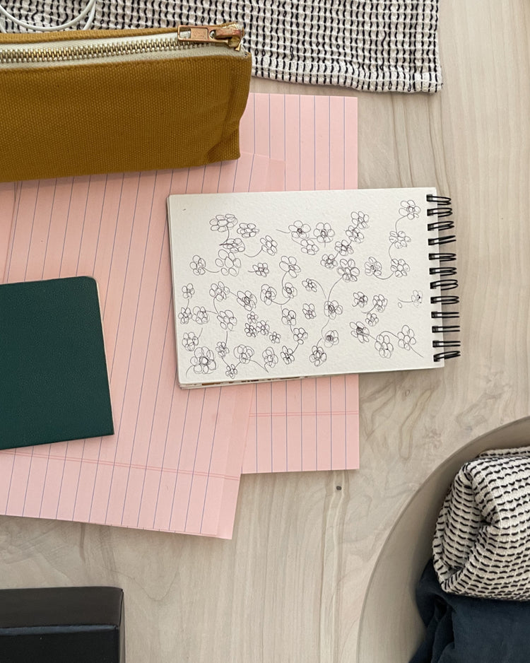 A styled flat-lay featuring a white spiral notebook with hand-drawn black floral sketches, resting on pink lined paper. The notebook is surrounded by other stationery items, including a mustard yellow pencil case, a dark green notebook, and a black-and-white patterned fabric, all arranged on a light wood surface.