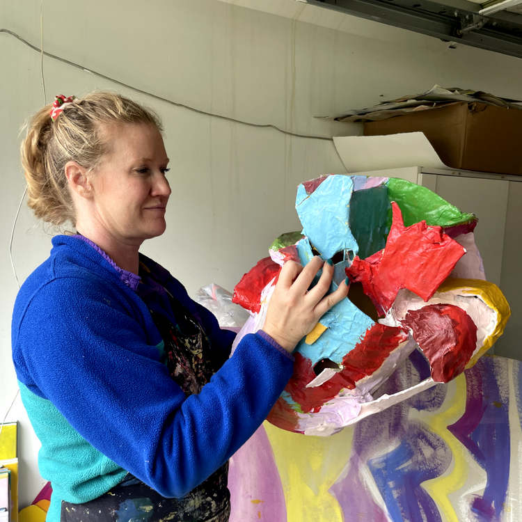 A woman in a blue fleece sweater holds a colorful abstract sculpture made of layered, textured paper or fabric pieces in red, blue, green, yellow, and white in an indoor studio space.