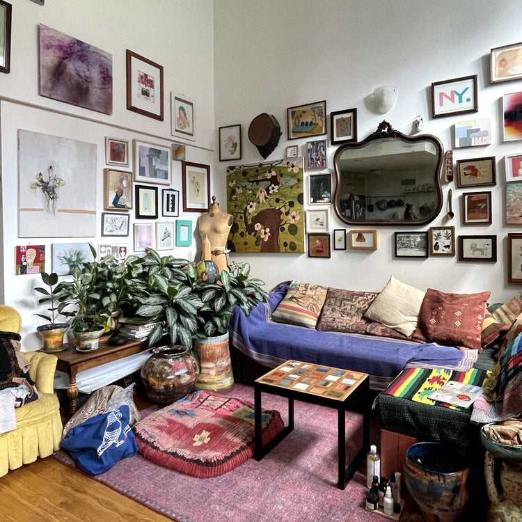A cozy, densely decorated living room corner with a blue sofa, pink rug, potted plants, framed wall art, a mirror, and a mannequin torso in the center.