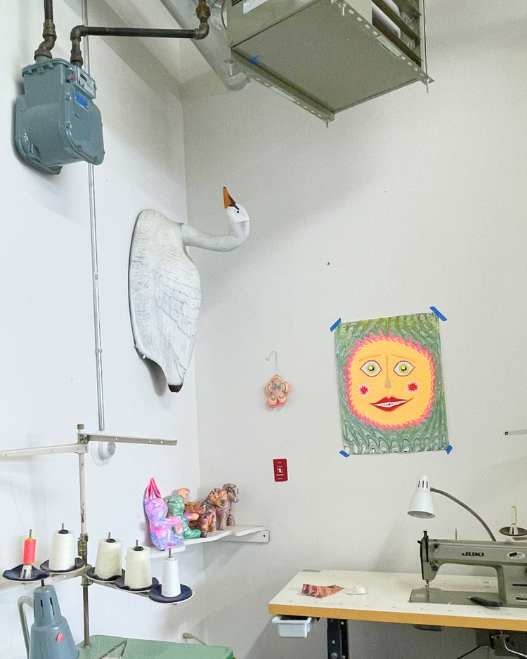 A cluttered sewing room corner with a white swan taxidermy mount on the wall, a colorful sun face tapestry, sewing machines, spools of thread, and industrial ceiling ductwork.