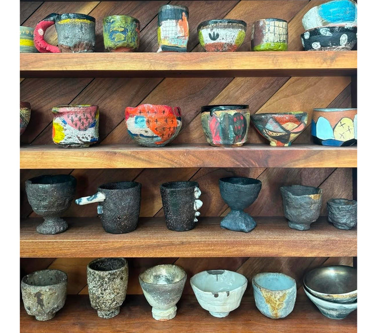 A collection of handmade, rustic ceramic mugs and cups displayed on three wooden shelves against a dark wood paneled background.