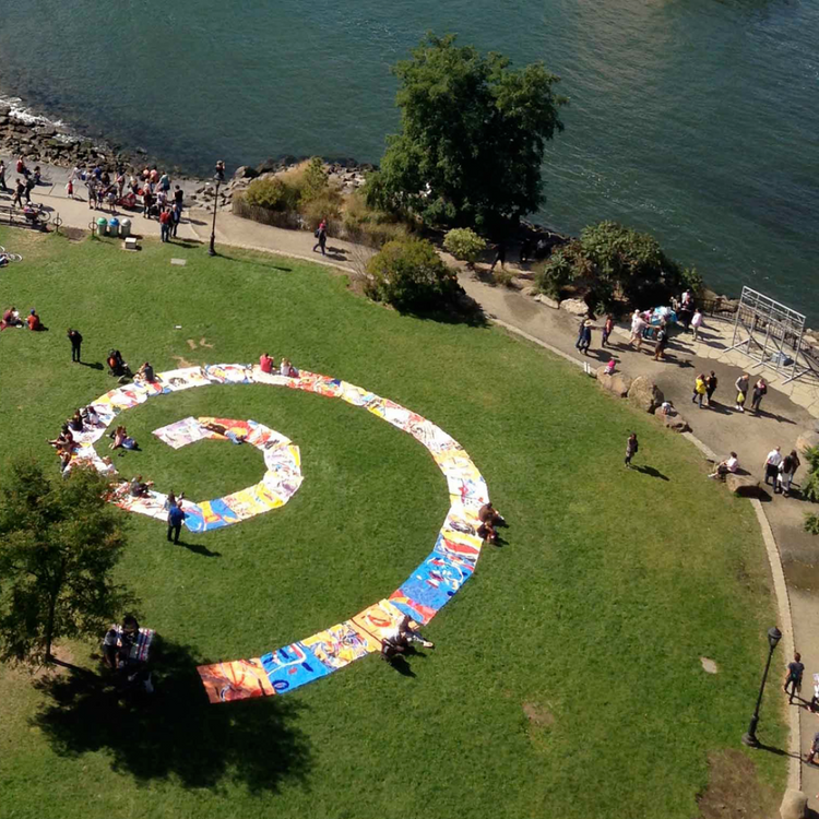 An aerial view of a large spiral arrangement of colorful blankets laid out on a green park lawn next to a body of water, with small groups of people standing and sitting around it.