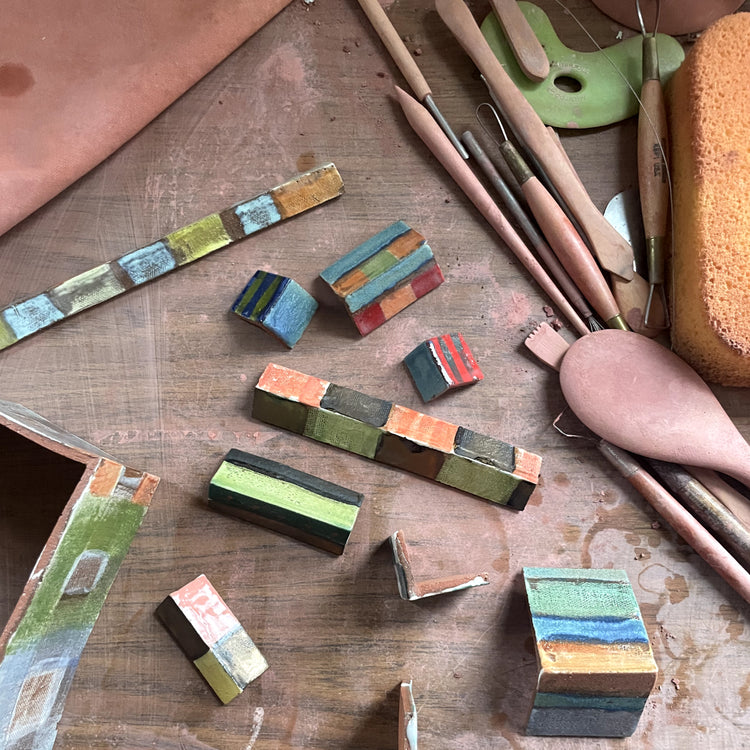A collection of hand-painted wooden blocks and strips with colorful geometric patterns, arranged on a rustic wooden workbench alongside pottery tools like wooden sculpting tools, a sponge, and a clay wheel.