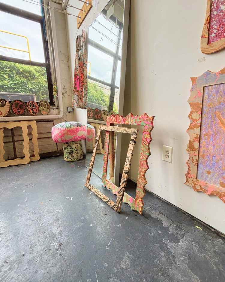 A room with a gray concrete floor and white walls, featuring several colorful, ornate decorative frames leaning against the wall and one mounted on the right wall. A pink-and-green patterned stool sits near the window on the left.