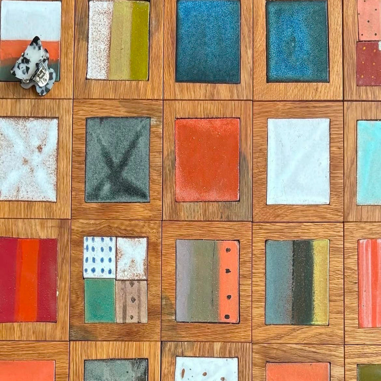 A close-up of a wooden board or tile featuring multiple square ceramic swatches in various colors and textures, including blue, orange, white, green, yellow, and patterned designs.