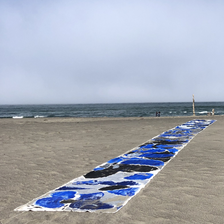 A long rectangular textile laid out on a sandy beach, featuring an abstract pattern of blue, black, and white brushstrokes. The background shows the ocean under a cloudy, overcast sky.