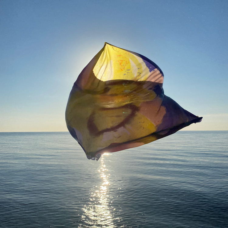 A translucent scarf with yellow, purple, and brown abstract patterns floating in the wind over a calm blue ocean under a bright sun.