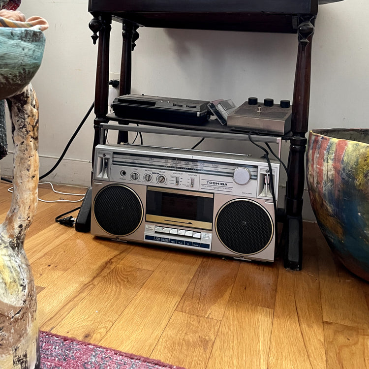 A vintage silver and gold Toshiba boombox radio sits on a wooden floor next to a dark wooden stand holding a turntable and cassette deck, with decorative ceramic planters on either side.
