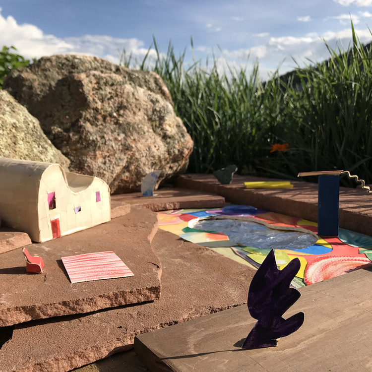 A miniature outdoor diorama scene with a small house, colorful painted stones, a painted pool, rocks, tall grass, and a blue sky in the background.