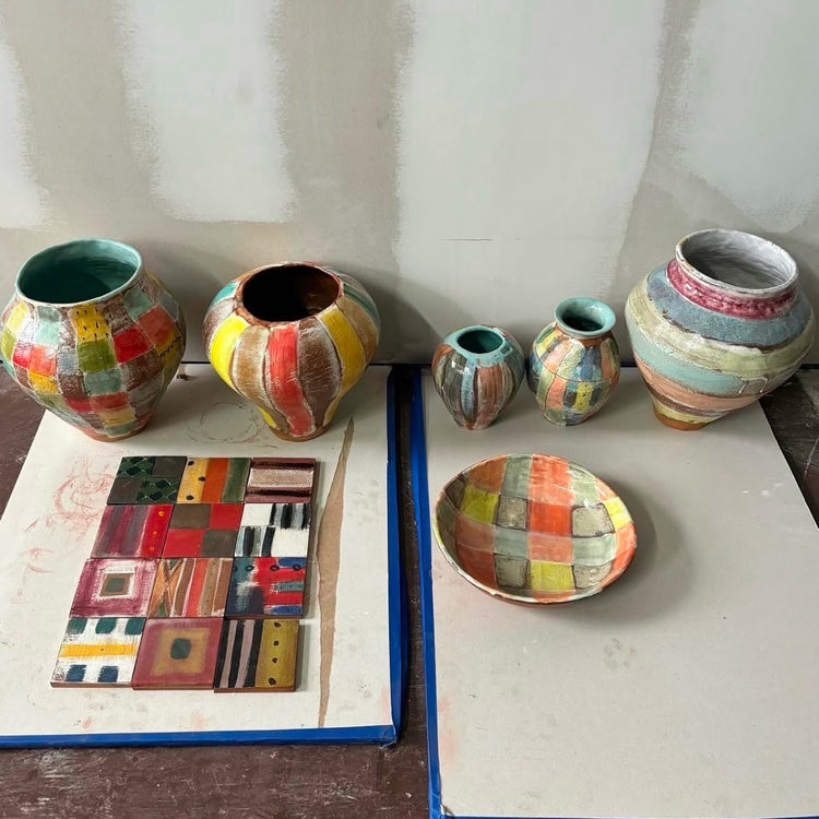 A collection of handmade ceramic pottery including vases, a bowl, and a square tile plaque, all decorated with colorful patchwork patterns in red, yellow, green, blue, and orange, displayed on a table against a gray wall.