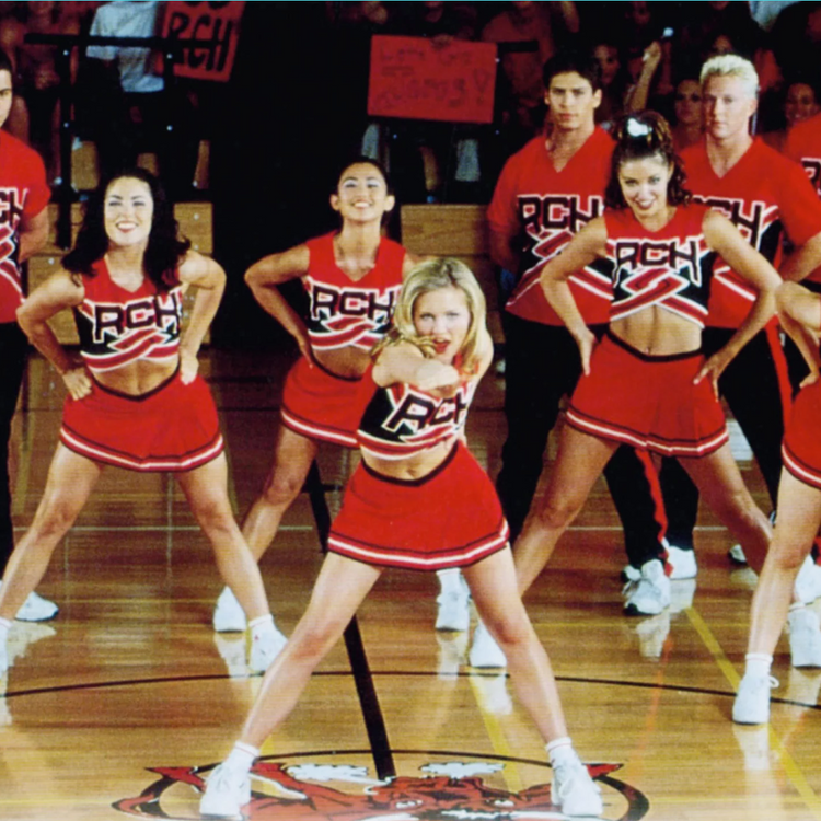 A group of cheerleaders in matching red, black, and white uniforms posing on a basketball court. The center cheerleader is in the foreground, with others behind her on a polished wooden court.