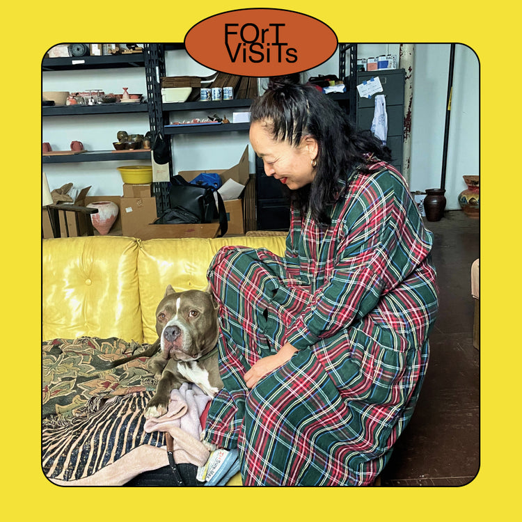 A woman in a green, red, and black plaid blanket pajama set sits on a yellow couch in a home setting, petting a brown and white pitbull-type dog. The photo is framed with a thick yellow border and an orange sticker at the top that reads 'FOR I VISITS'.