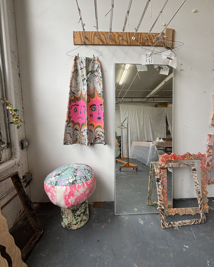 A styled interior scene showing a tall floor mirror, a pink and multicolor patterned curtain, a round upholstered stool, and several distressed picture frames in a studio or workshop setting.