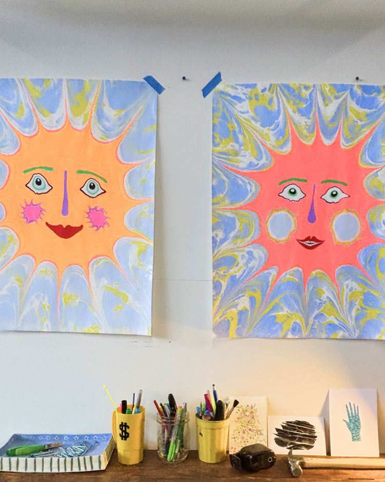 Two colorful sun paintings hanging on a white wall above a desk with art supplies. The left painting has a yellow sun with blue and white marbled rays, and the right painting has a pink sun with blue, yellow, and white marbled rays.