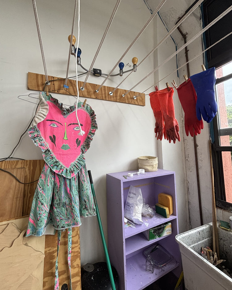 A pink heart-shaped dress with a printed pattern hangs on a clothesline in a room. Red and blue gloves are hanging on the right side, with a purple shelf and other household items visible in the background.