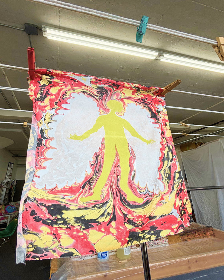 A large fabric art print featuring a stylized human figure silhouette in yellow, surrounded by red, white, and black marbled patterns, hanging from wooden clips on a drying rack in an indoor studio space.